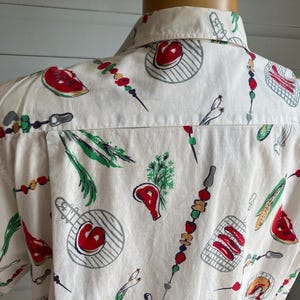 May include: Off-white short-sleeved shirt with a repeating pattern of barbecue-themed illustrations. The design includes red watermelon slices, grilled meat, skewers, and grilling utensils. The shirt is made of a lightweight fabric.