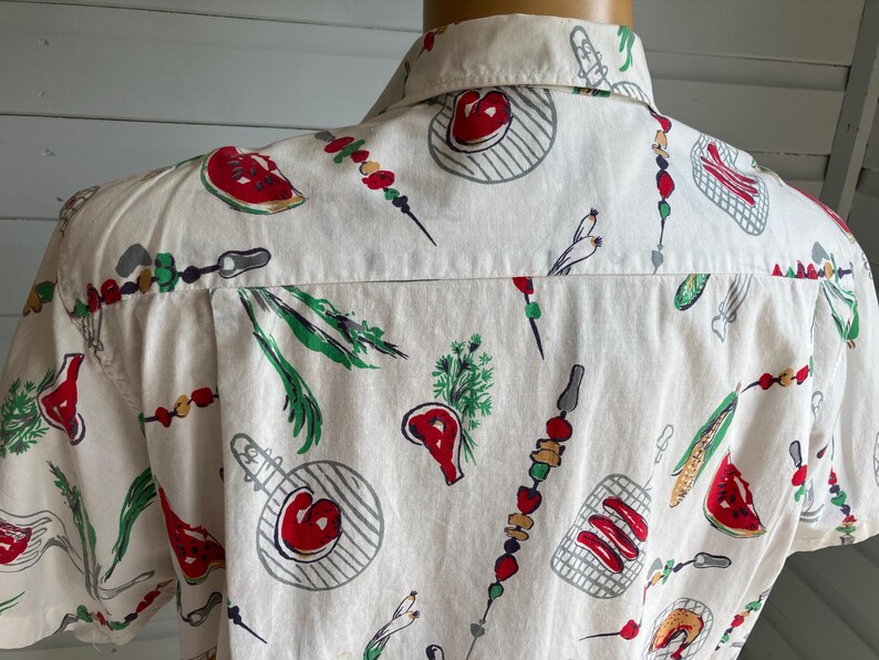 May include: Off-white short-sleeved shirt with a repeating pattern of barbecue-themed illustrations. The design includes red watermelon slices, grilled meat, skewers, and grilling utensils. The shirt is made of a lightweight fabric.