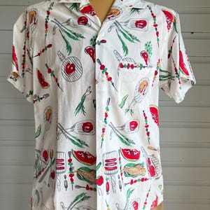 May include: White short-sleeve shirt with a repeating pattern of barbecue-themed illustrations. The design includes images of grilled meats, vegetables, and grilling utensils in red, green, and gray. The shirt has a classic collar and a single chest pocket.