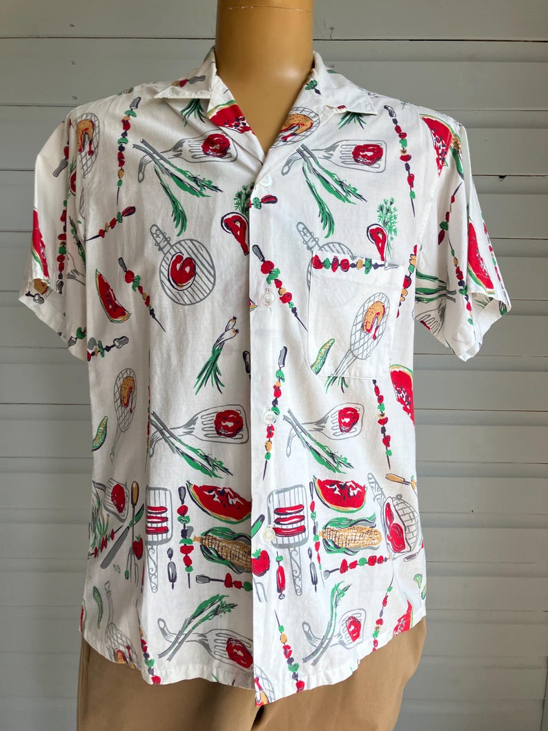 May include: White short-sleeve shirt with a repeating pattern of barbecue-themed illustrations. The design includes images of grilled meats, vegetables, and grilling utensils in red, green, and gray. The shirt has a classic collar and a single chest pocket.
