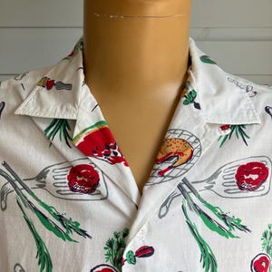 May include: White button-down shirt with a colorful food-themed print. The pattern includes images of grilled meats, vegetables, and cooking utensils in red, green, and gray. The shirt has a classic collar and short sleeves.