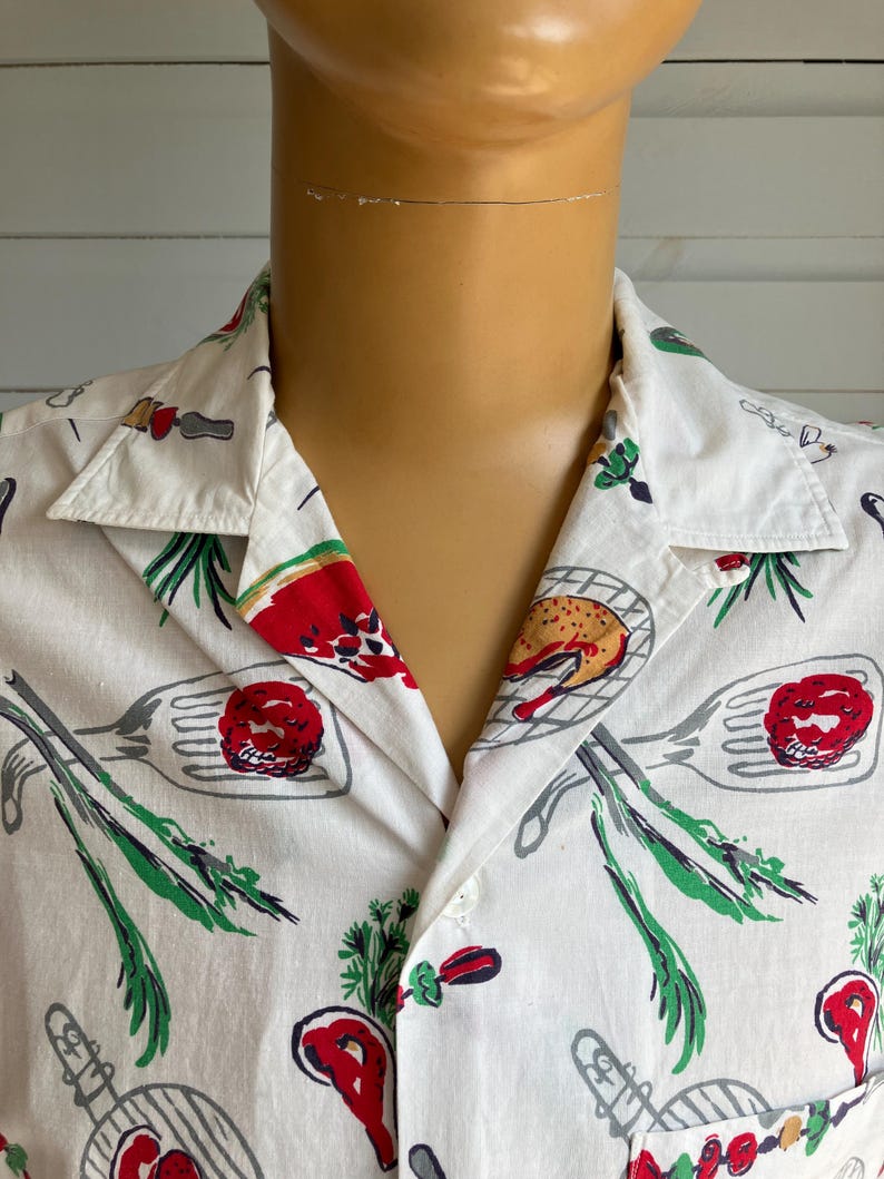 May include: White button-down shirt with a colorful food-themed print. The pattern includes images of grilled meats, vegetables, and cooking utensils in red, green, and gray. The shirt has a classic collar and short sleeves.