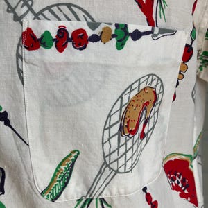 May include: White button-up shirt with a pocket, featuring a colorful barbecue-themed print. The design includes illustrations of grilled food, skewers, and grilling tools in red, green, brown, and gray. The shirt's fabric appears to be a lightweight linen or cotton.
