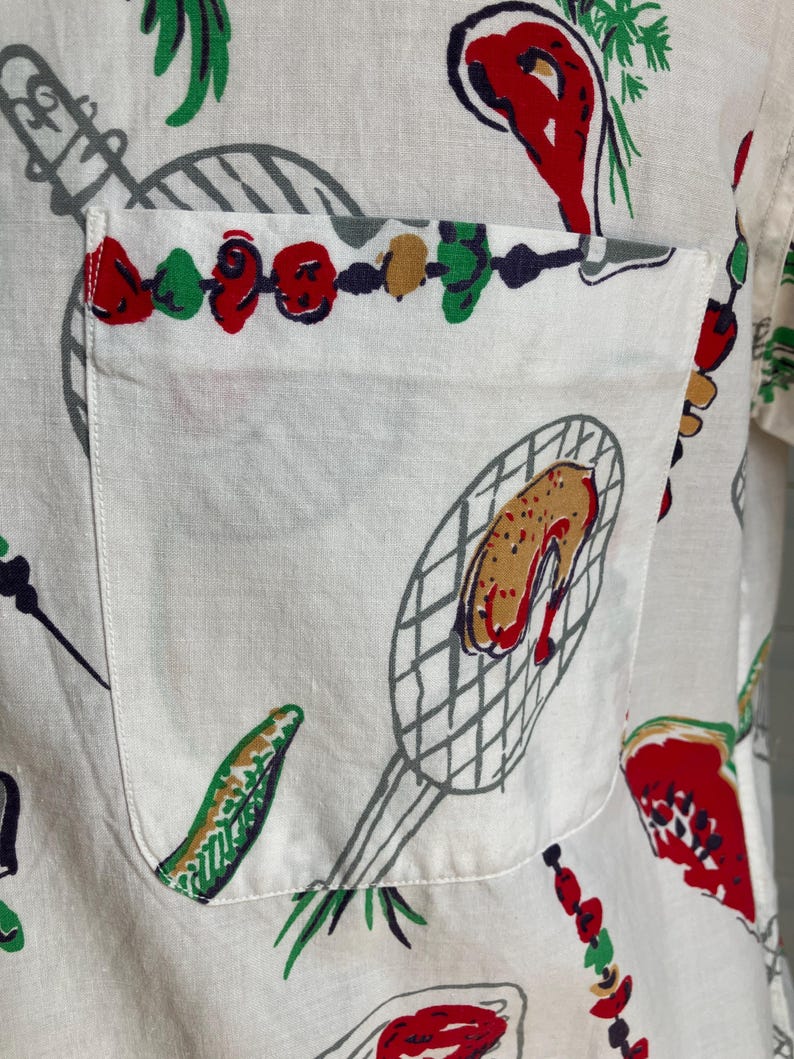 May include: White button-up shirt with a pocket, featuring a colorful barbecue-themed print. The design includes illustrations of grilled food, skewers, and grilling tools in red, green, brown, and gray. The shirt's fabric appears to be a lightweight linen or cotton.
