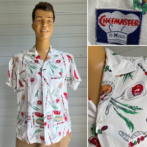 May include: White short-sleeve shirt with a print of grilling tools, vegetables, and food items. The shirt has a camp collar and a chest pocket. The tag reads "Chefmaster 15 M 15 1/2".