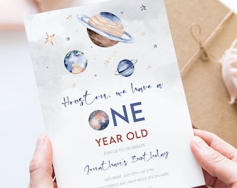 May include: A white invitation card with a blue and brown watercolor illustration of planets and stars. The text on the card reads "Houston, we have a ONE YEAR OLD. Join us to celebrate Jonathan's Birthday. Saturday, July 1st | 4:30 PM | 123 Butterfly Ave, Belle Ville, 01234 5678. Registered at Amazon."