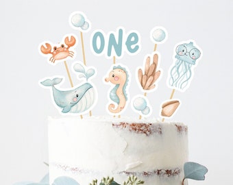 May include: A white cake decorated with blue and orange sea-themed decorations. The cake features a whale, a seahorse, a crab, a jellyfish, and a seashell. The cake also has the word "ONE" on it. The cake is on a white cake stand with green leaves around it.