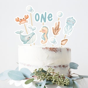 May include: A white cake with blue and orange decorations. The cake is decorated with a whale, a seahorse, a crab, a jellyfish, and a seashell. The cake also has the word "ONE" on it. The cake is on a white cake stand with green leaves around it.