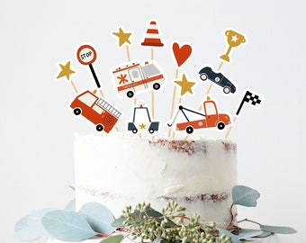May include: A white cake with red, blue, and yellow vehicle themed cake toppers. The cake is on a white cake stand with green leaves around the base.