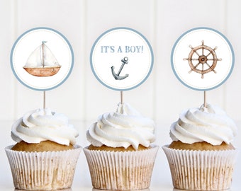 Puede incluir: Tres cupcakes con glaseado blanco cubiertos con círculos de papel redondos. Los círculos tienen bordes azules y presentan imágenes con temática náutica: un velero, un ancla con el texto "¡Es un niño!" y un timón.