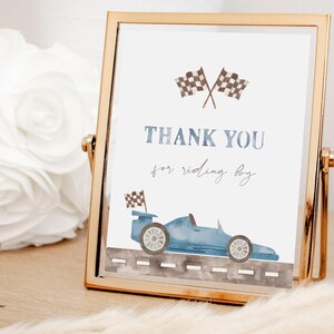 May include: A gold-framed sign with a blue race car and checkered flags. The sign says "Thank you for riding by".
