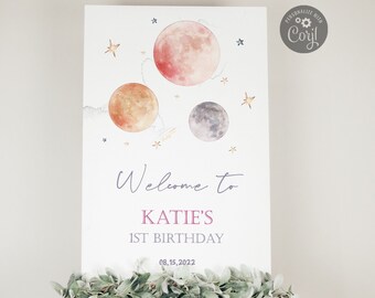May include: Welcome to Katie's 1st Birthday party sign with a watercolor illustration of planets and stars. The sign is on a wooden easel.