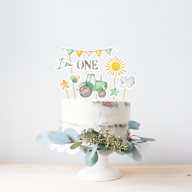 May include: A white cake with a green tractor, sun, clouds, and stars on top. The cake is decorated with a banner that says "I'm ONE".