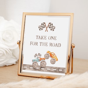 May include: A gold-framed sign with a white background and the text "Take one for the road". The sign features a watercolor illustration of two monster trucks with checkered flags.