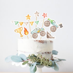 May include: A white cake with a blue monster truck, a trophy, a flame, and stars on top. The cake is decorated with a green, orange, and white banner.