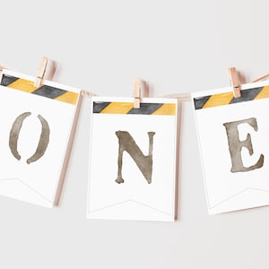 May include: A white banner with black and yellow stripes hangs with clothespins. The banner spells out "ONE" in large, gray, watercolor letters.