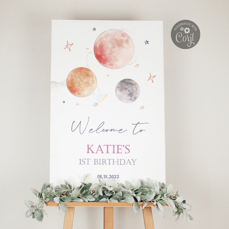 May include: Welcome to Katie's 1st Birthday party sign with a watercolor illustration of planets and stars. The sign is on a wooden easel.