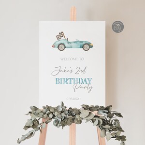 May include: A blue and white welcome sign for a birthday party. The sign features a watercolor illustration of a blue race car with a checkered flag. The text on the sign reads "Welcome to Jake's 2nd Birthday Party 07.15.2022".