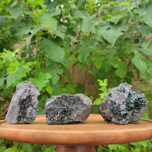 May include: Three green and brown rough-hewn crystals with a sparkly, iridescent finish. The crystals are arranged on a wooden display stand.