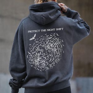 Puede incluir: Una sudadera con capucha de color gris jaspeado oscuro con un gráfico blanco en la espalda. El gráfico muestra un enjambre circular de murciélagos con el texto PROTECT THE NIGHT SHIFT impreso en letras mayúsculas blancas sobre la ilustración.