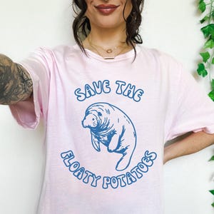 Puede incluir: Camiseta rosa con un gráfico azul de un manatí y el texto "Save the Floaty Potatoes".