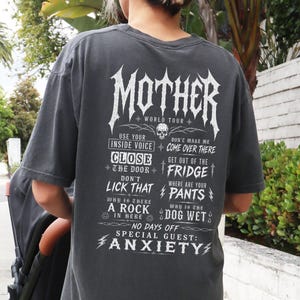 May include: A charcoal gray short-sleeve t-shirt featuring white graphic text on the back that reads MOTHER WORLD TOUR, followed by a list of phrases including USE YOUR INSIDE VOICE, DON'T LICK THAT, and SPECIAL GUEST: ANXIETY.