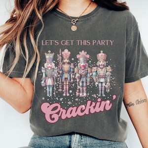 Puede incluir: Una camiseta gris con un gráfico rosa de soldados cascanueces y el texto "Let's get this party crackin'"