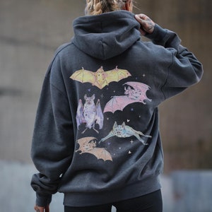 May include: A dark gray hooded sweatshirt with a graphic print of five bats in various colors and poses. The bats are outlined in black and have a watercolor effect. The sweatshirt has a relaxed fit and a drawstring hood.