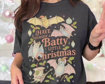 May include: Dark gray t-shirt with a festive design featuring bats, holly, and ornaments. The text reads "Have Yourself a Batty Little Christmas" in a whimsical font. The bats are in shades of yellow, pink, and gray.