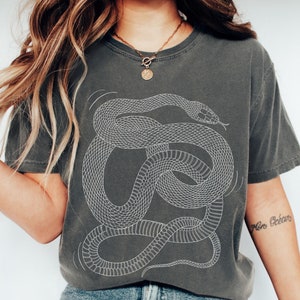 May include: A dark grey t-shirt featuring a white outline of a snake design. The snake is curled up in a circle with its head facing the right side of the shirt. The shirt has a relaxed fit and a cropped length.