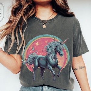 May include: A dark gray t-shirt with a graphic of a blue unicorn standing in front of a rainbow and pink background with stars. The unicorn has a gold horn and a gold star on its side.