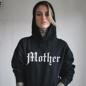 May include: Black hooded sweatshirt with the word "Mother" in a gothic font across the chest. The sweatshirt has a drawstring hood and a front pocket. The word "black" is in the bottom right corner.