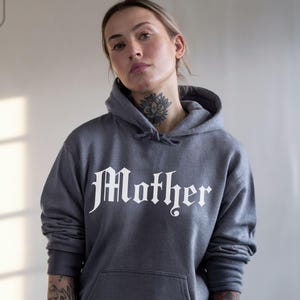 May include: Dark heather gray hoodie with the word "Mother" in a gothic font across the chest. The hoodie has a drawstring hood and a front pocket. The image also shows a small logo in the upper left corner and the words "dark heather" in the bottom right corner.