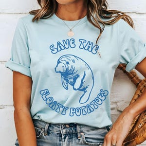 Puede incluir: Camiseta azul claro con una ilustración de manatí azul y el texto "Save the Floaty Potatoes". La camiseta tiene mangas enrolladas y está hecha de un material suave. La camiseta se usa con jeans azules.