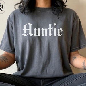 May include: A dark gray t-shirt with the word "Auntie" printed in white, gothic-style lettering. The shirt has short sleeves and a relaxed fit. The image shows the shirt on a person sitting down.