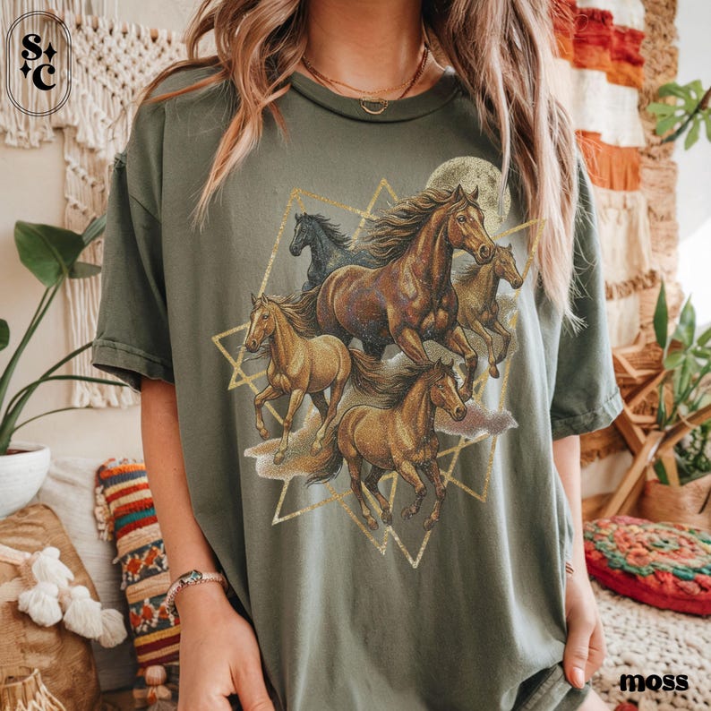 May include: Moss green t-shirt with a graphic of running horses in brown and gold, set against a full moon and geometric shapes. The shirt has short sleeves and a relaxed fit.