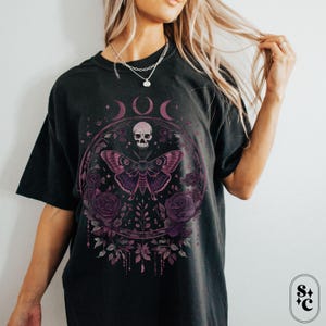 K&ouml;nnte beinhalten: Schwarzes T-Shirt mit einem grafischen Druck eines Sch&auml;dels, eines Nachtfalters und Blumen. Die Grafik ist in Rosa- und Lilat&ouml;nen gehalten. Das Shirt hat einen Used-Look.