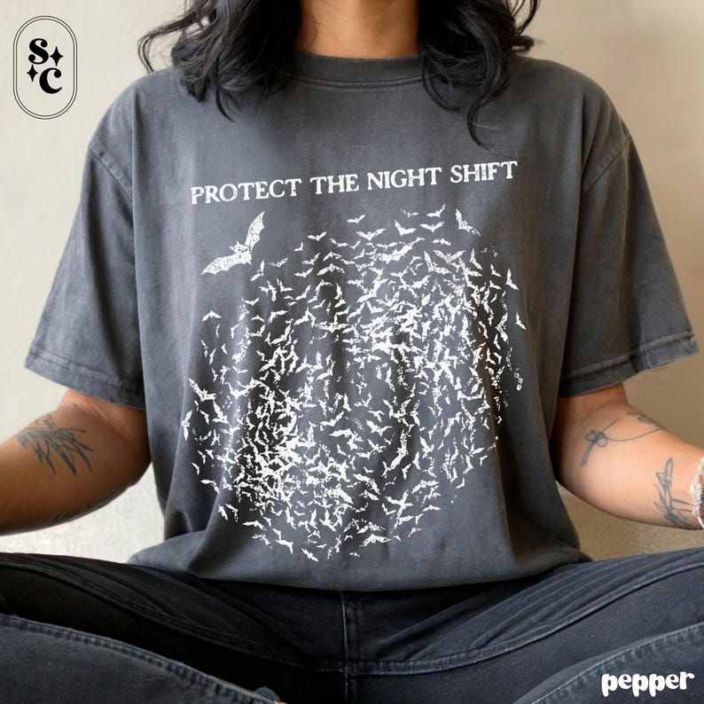 May include: Dark gray t-shirt with the text "PROTECT THE NIGHT SHIFT" above a graphic of a swarm of bats. The bats are white and clustered together, with a single bat flying above the swarm. The shirt is a casual style.
