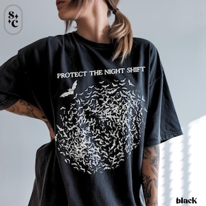 May include: Black t-shirt with the text "PROTECT THE NIGHT SHIFT" above a cluster of white bat illustrations. The word "black" is printed at the bottom right corner. The shirt is a dark color.