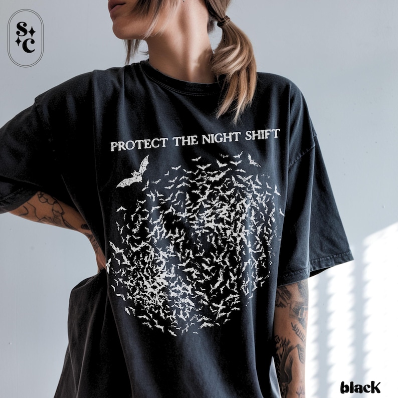 May include: Black t-shirt with the text "PROTECT THE NIGHT SHIFT" above a cluster of white bat illustrations. The word "black" is printed at the bottom right corner. The shirt is a dark color.
