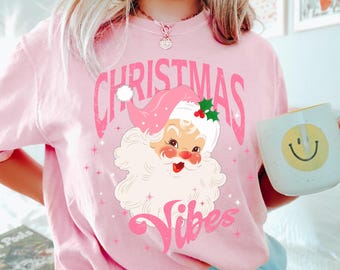 May include: A pink Christmas-themed t-shirt with a vintage Santa Claus graphic. The design features the words "Christmas Vibes" in a pink font. The Santa Claus illustration has a pink hat and a white beard, evoking a retro style.