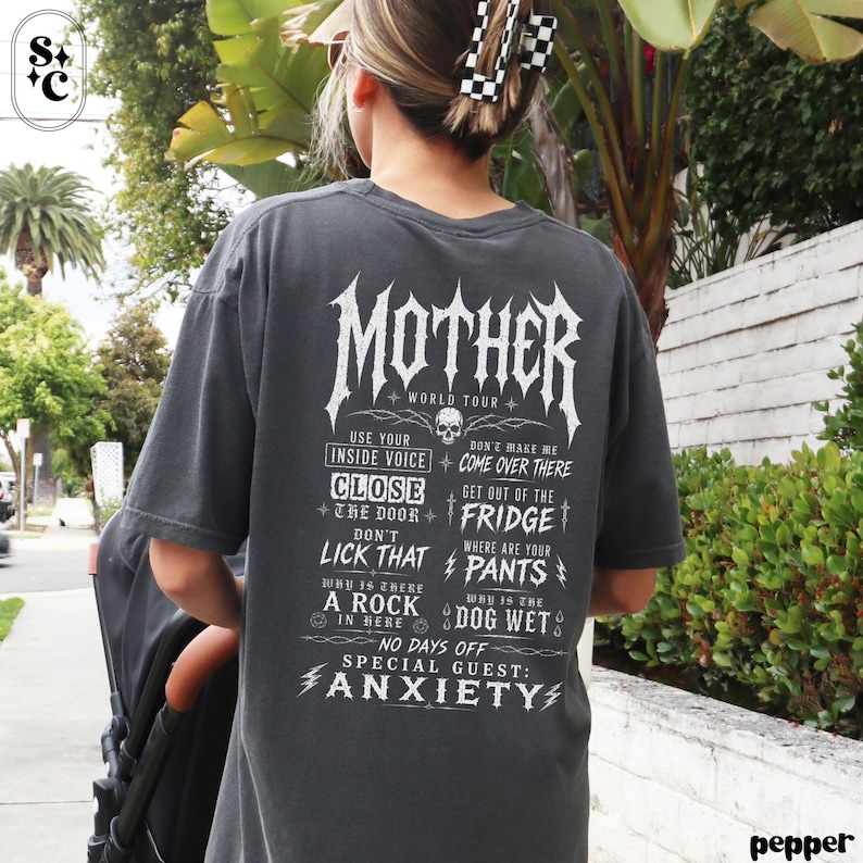 May include: A charcoal gray short-sleeve t-shirt featuring white graphic text on the back that reads MOTHER WORLD TOUR, followed by a list of phrases including USE YOUR INSIDE VOICE, DON'T LICK THAT, and SPECIAL GUEST: ANXIETY.