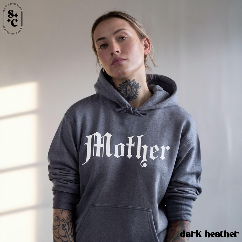 May include: Dark heather gray hoodie with the word "Mother" in a gothic font across the chest. The hoodie has a drawstring hood and a front pocket. The image also shows a small logo in the upper left corner and the words "dark heather" in the bottom right corner.