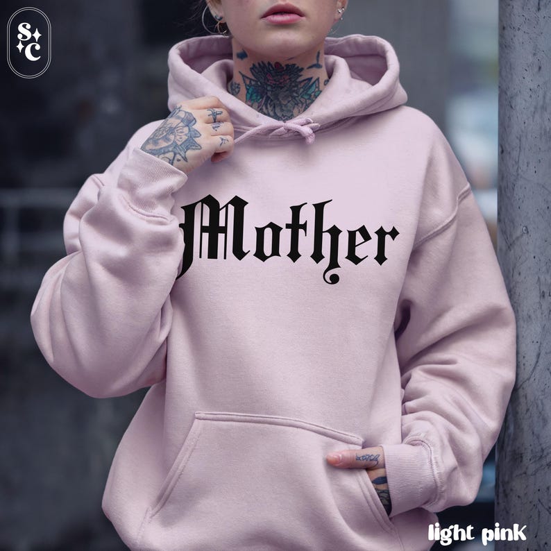 May include: Light pink hoodie with the word "Mother" in a gothic font across the chest. The hoodie has a kangaroo pocket and a hood with a drawstring. The words "light pink" are at the bottom right.