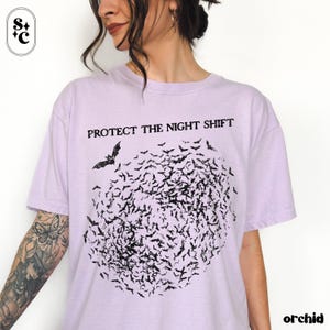 May include: A light purple t-shirt with the text "PROTECT THE NIGHT SHIFT" above a flock of black bats. The word "orchid" is printed in the bottom right corner. The shirt is a casual, comfortable style.