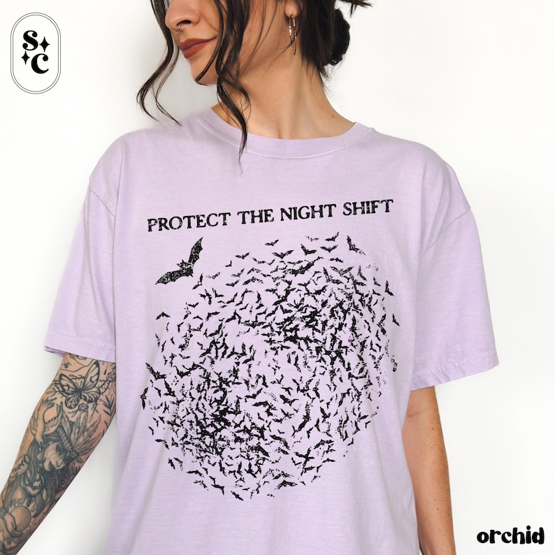 May include: A light purple t-shirt with the text "PROTECT THE NIGHT SHIFT" above a flock of black bats. The word "orchid" is printed in the bottom right corner. The shirt is a casual, comfortable style.