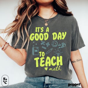 Op de afbeelding: Donkergrijs t-shirt met de tekst "IT'S A GOOD DAY TO TEACH math" in neon groen en lichtblauw. Het shirt heeft wiskundegerelateerde afbeeldingen zoals een notitieboek, potlood en gloeilamp. Het woord "pepper" staat rechtsonder.
