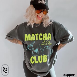 Puede incluir: Camiseta gris oscuro con un dise&ntilde;o "Matcha Club" en verde ne&oacute;n. El dise&ntilde;o incluye una taza de t&eacute;, un batidor, un cuenco y una tetera. Las palabras "all day, every day" tambi&eacute;n est&aacute;n incluidas.
