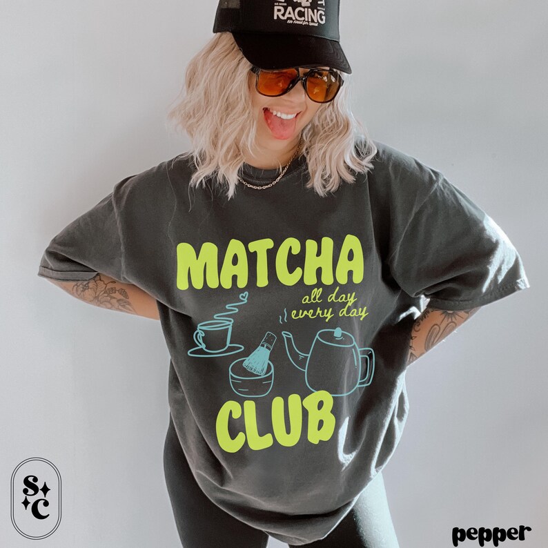 Puede incluir: Camiseta gris oscuro con un dise&ntilde;o "Matcha Club" en verde ne&oacute;n. El dise&ntilde;o incluye una taza de t&eacute;, un batidor, un cuenco y una tetera. Las palabras "all day, every day" tambi&eacute;n est&aacute;n incluidas.