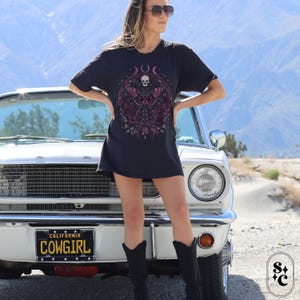 K&ouml;nnte beinhalten: Ein schwarzes T-Shirt mit einem Motiv aus einem Totenkopf und einem Schmetterling mit rosa und violetten Akzenten. Der Text "DOC" befindet sich &uuml;ber dem Motiv. Das Model tr&auml;gt das Shirt und steht vor einem wei&szlig;en Auto mit einem Nummernschild, auf dem "CALIFORNIA COWGIRL" steht.